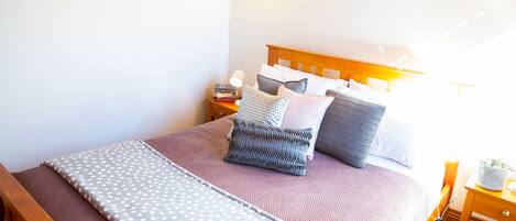 Una cama bien hecha con cubrecama y almohadas estampadas, una mesita de noche con lámpara y un mueble auxiliar con libros.