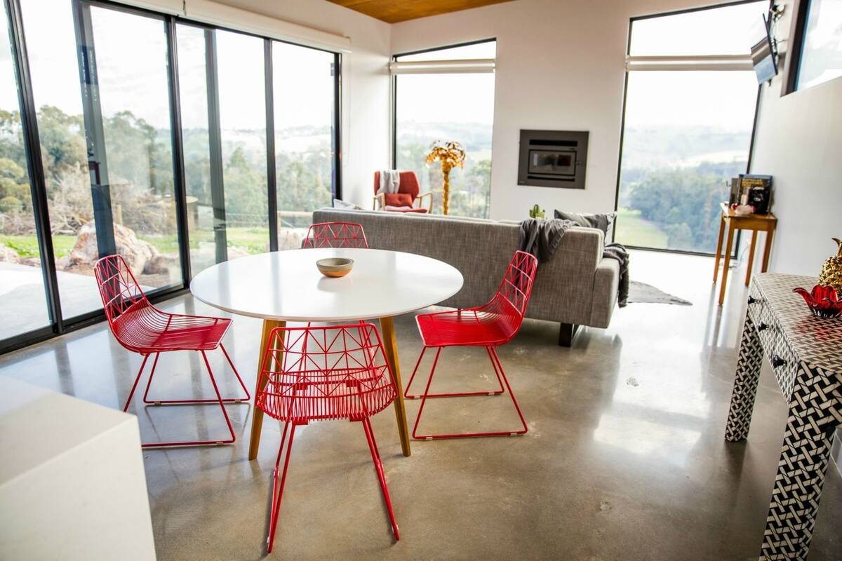 Uma sala de jantar moderna com uma mesa redonda e cadeiras vermelhas, amplas janelas com vista para o exterior, e um espaço de estar acolhedor com um sofá e poltronas.