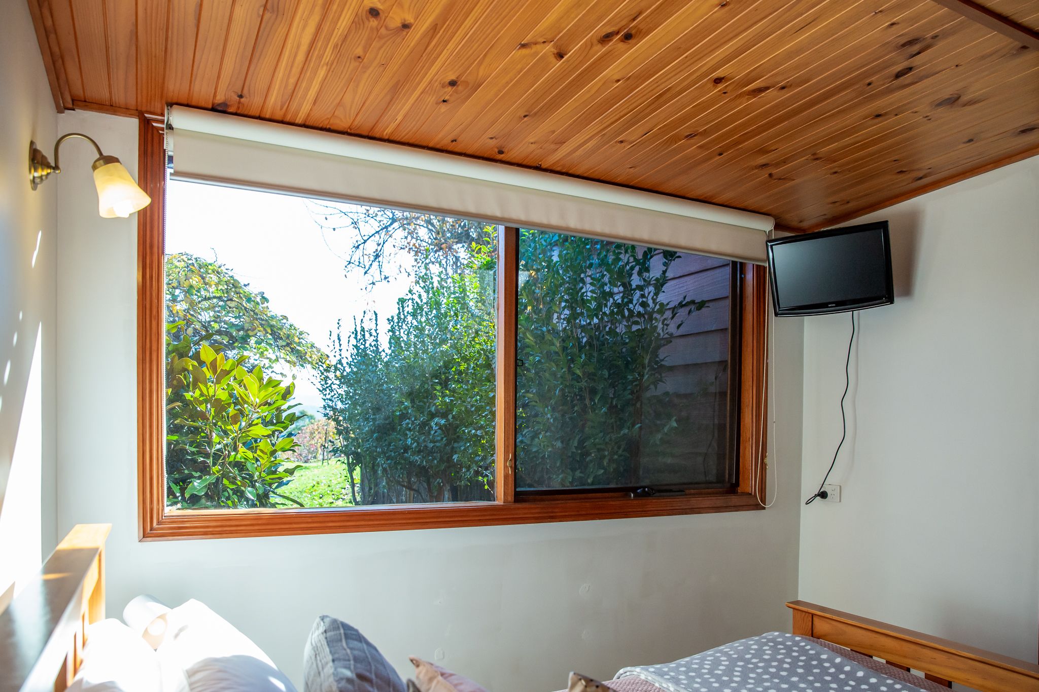 A bedroom with a large window, a bed, a bedside table, a lamp, and a TV mounted on the wall.