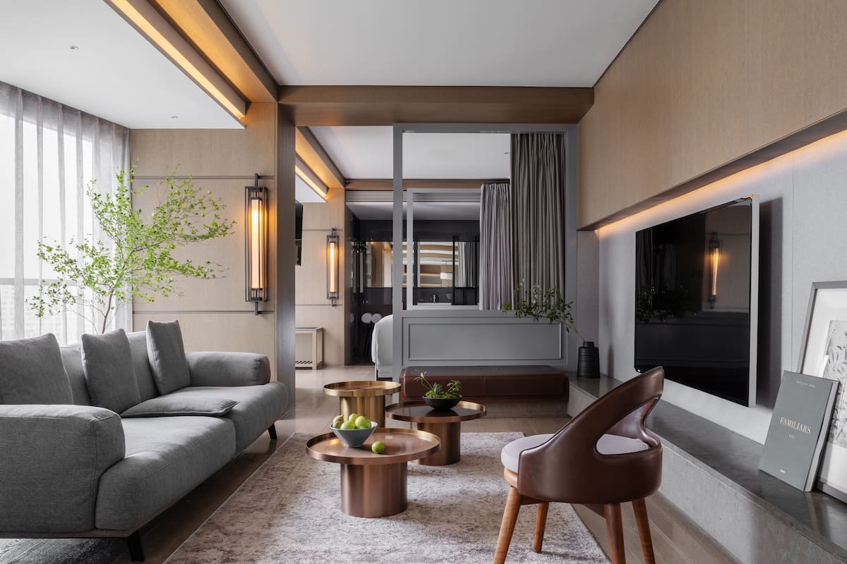 Un salon moderne avec un canapé gris, un fauteuil en cuir marron et une table basse sur laquelle on peut voir des pommes. Il y a un téléviseur accroché au mur et une fenêtre avec des rideaux.