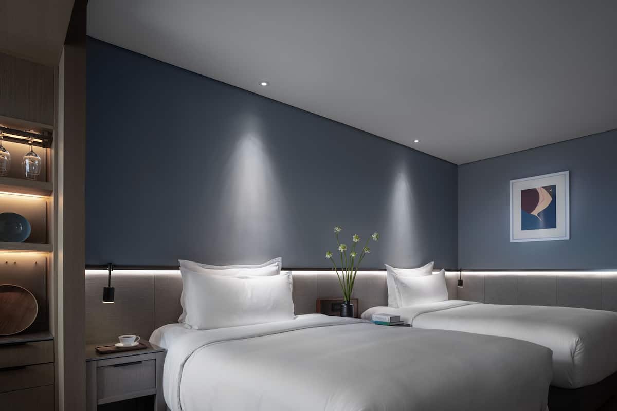 Une chambre d’hôtel moderne avec deux lits, une table de chevet avec une tasse, un vase avec des fleurs et un tableau encadré au mur.