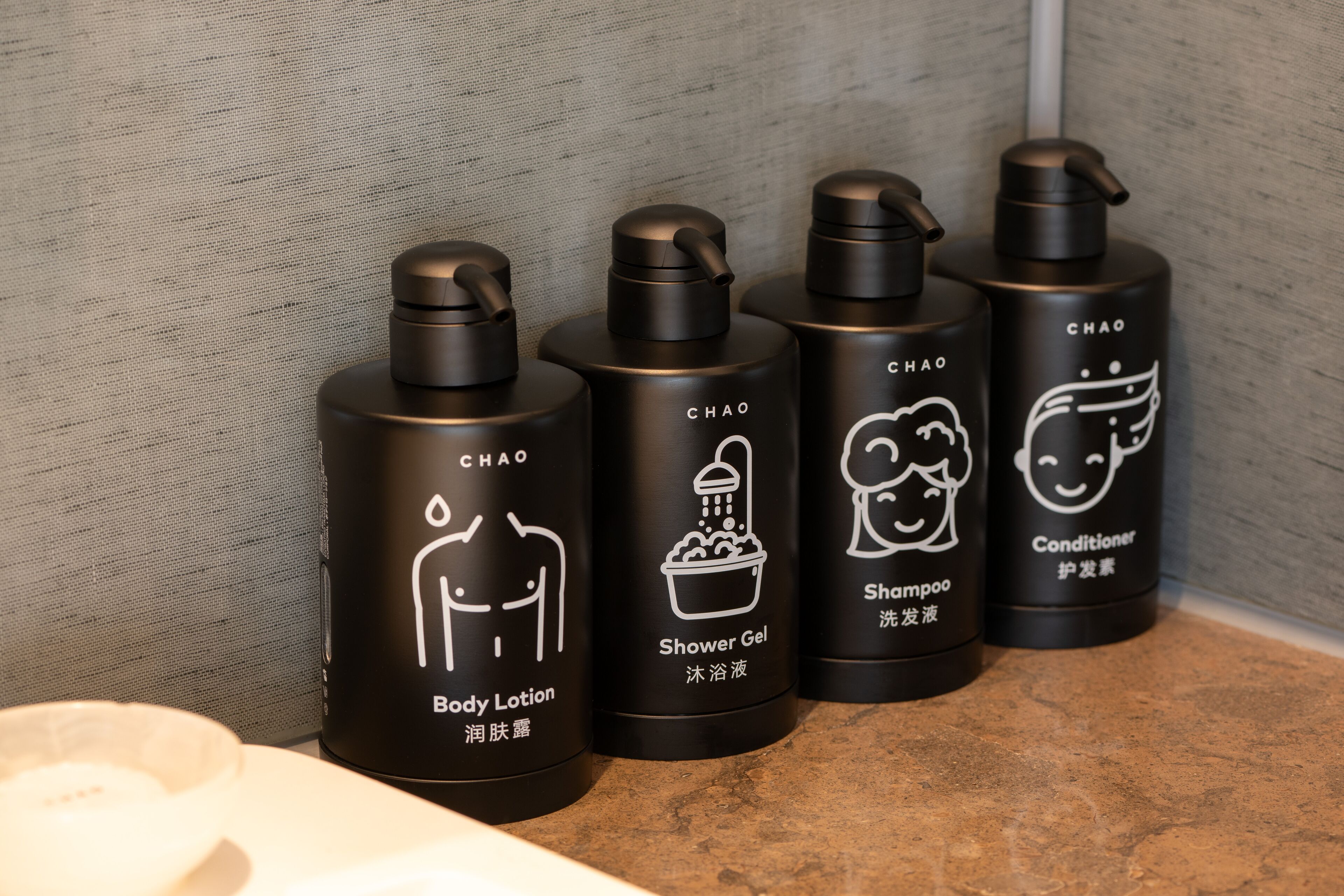 Four black bottles with white text and icons labeled as body lotion, shower gel, shampoo, and conditioner.