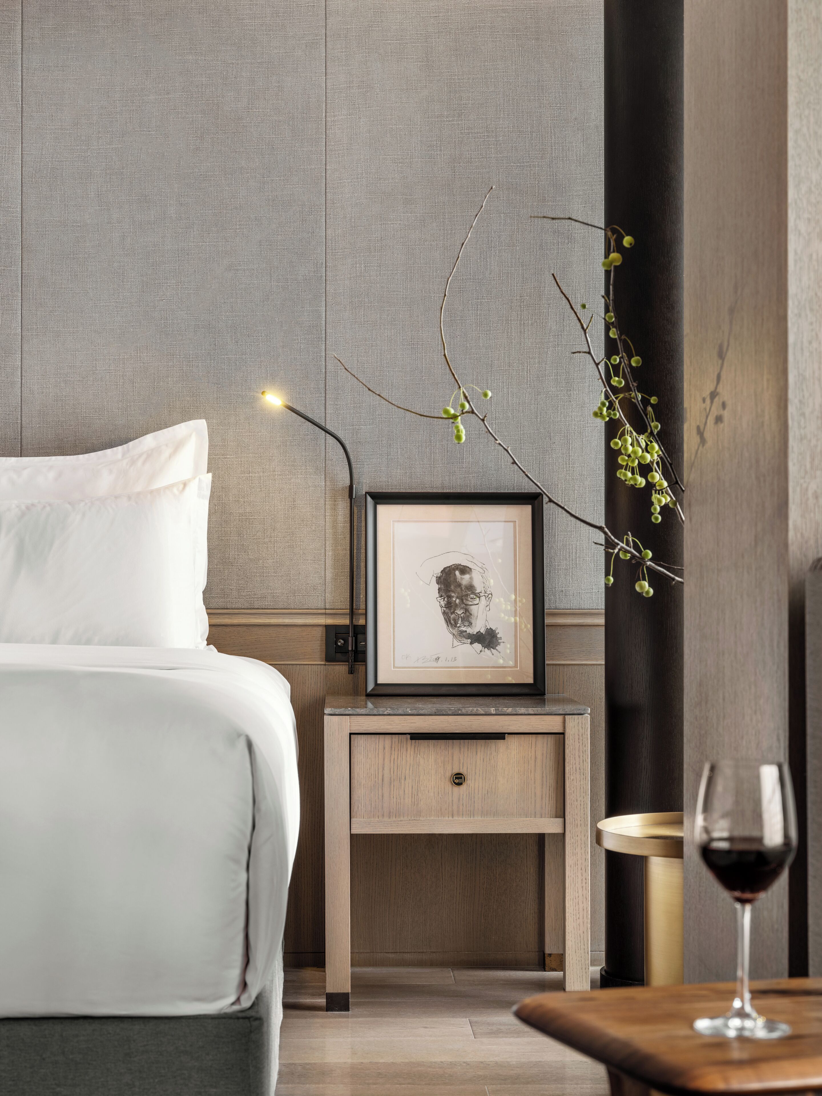 Ein modernes Hotelzimmer mit einem ordentlich bezogenen Bett, einem Nachttisch mit einem gerahmten Kunstwerk, einer kleinen Vase mit einem Zweig und einem Weinglas auf einem Holztisch.