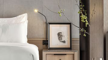 Ein modernes Hotelzimmer mit einem ordentlich bezogenen Bett, einem Nachttisch mit einem gerahmten Kunstwerk, einer kleinen Vase mit einem Zweig und einem Weinglas auf einem Holztisch.