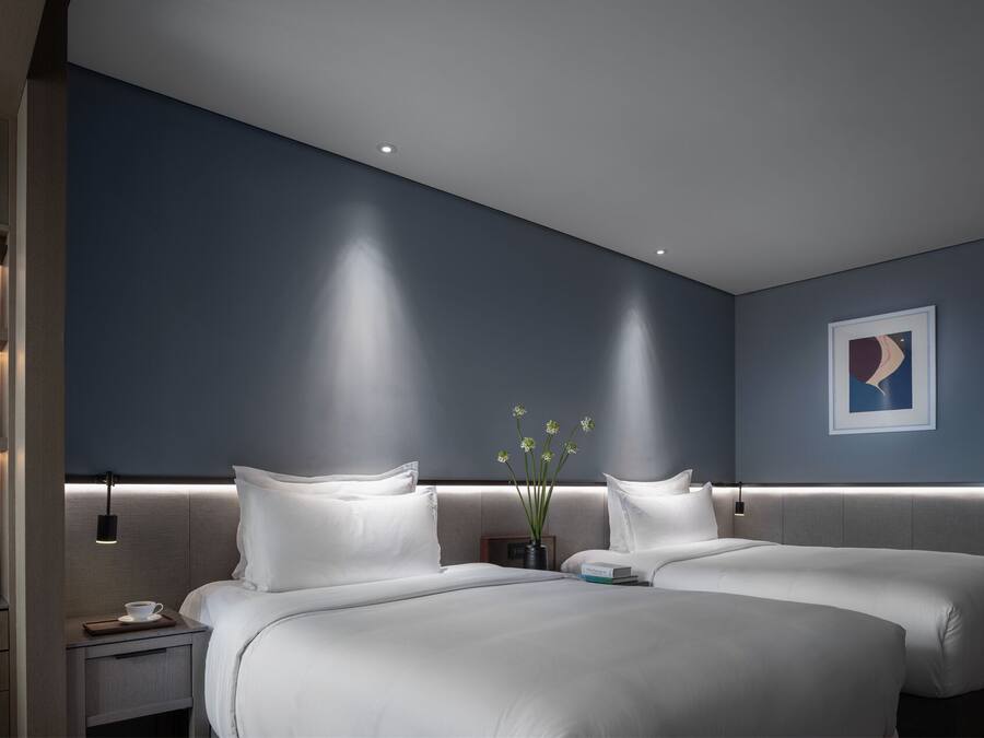 A modern hotel room with a large bed, bedside tables, a nightstand, and a wall-mounted shelf with decorative items.