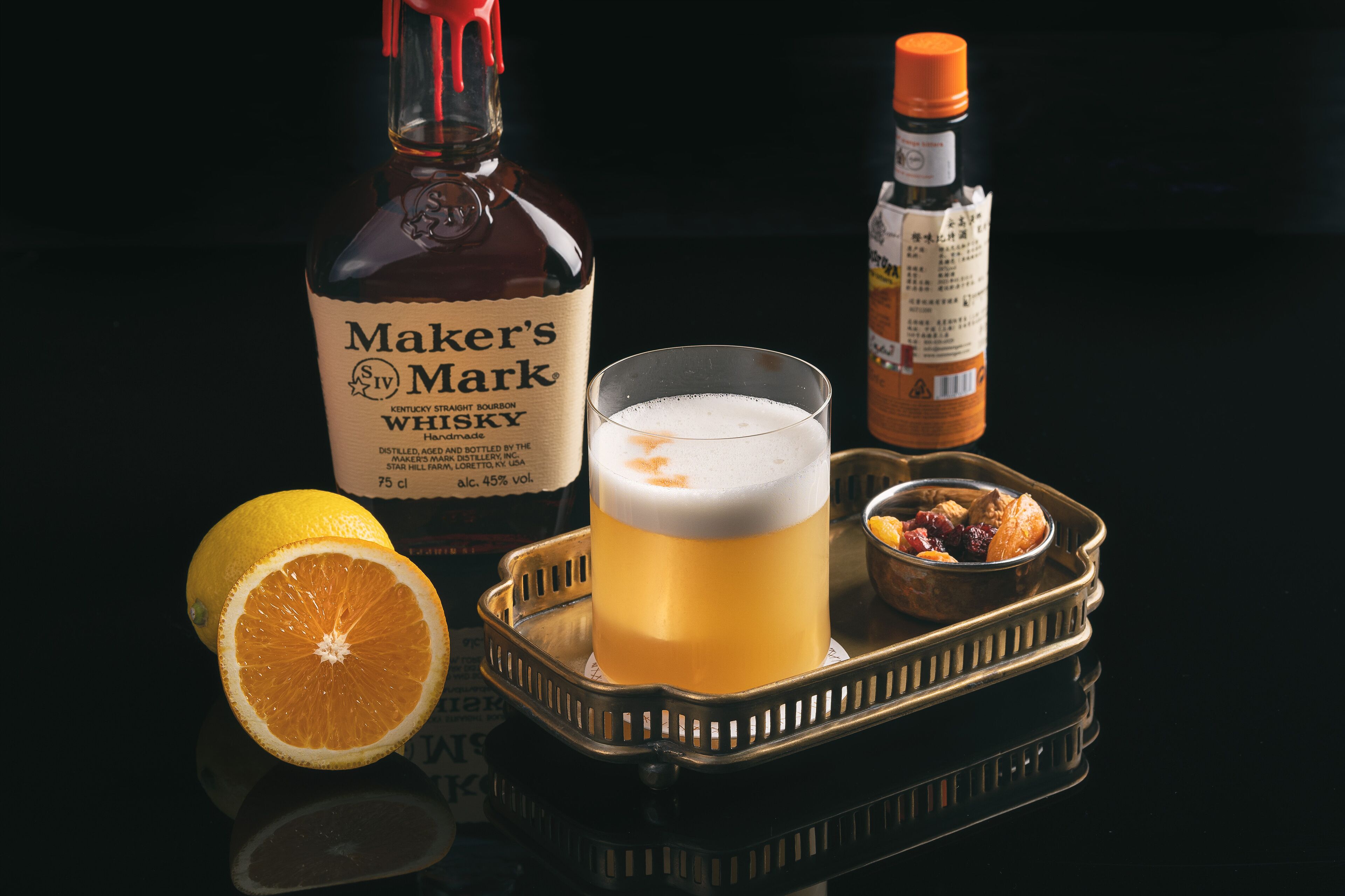 A tray with a glass of whiskey, a bottle of Maker's Mark whiskey, a bottle of hot sauce, a halved orange, and a small bowl of nuts.