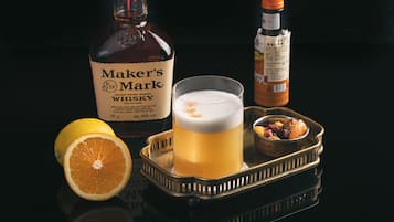 A tray with a glass of whiskey, a bottle of Maker's Mark whiskey, a bottle of hot sauce, a halved orange, and a small bowl of nuts.