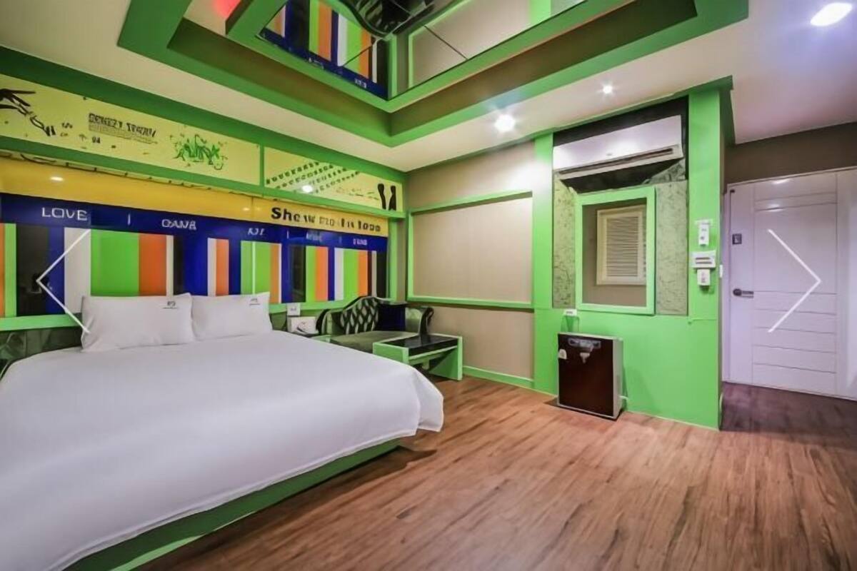 A hotel room with a bed, a desk, a chair, a trash bin, and a door.