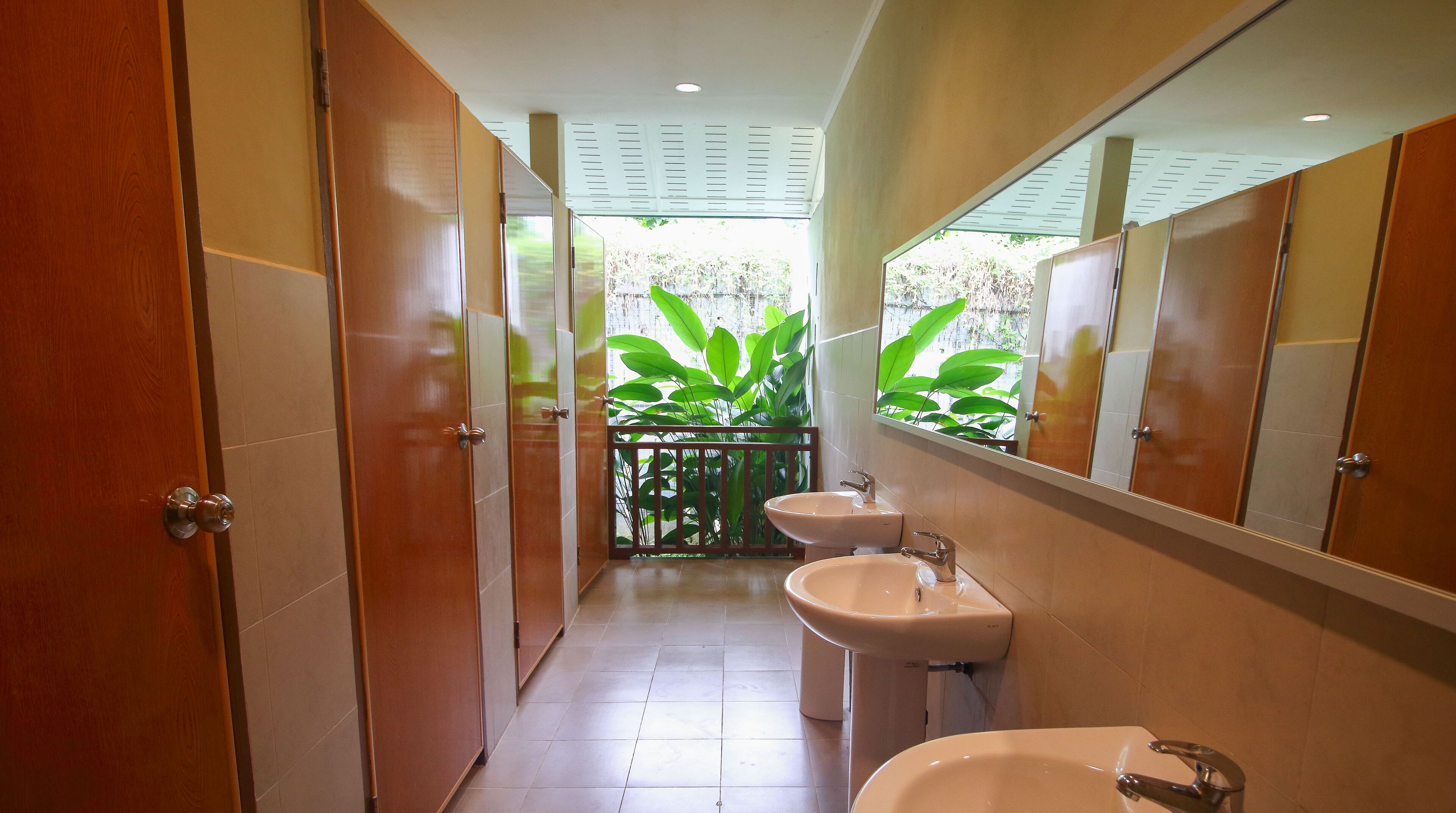 A narrow hallway with wooden doors and a mirrored wall, leading to a bathroom with two sinks and a view of greenery outside.