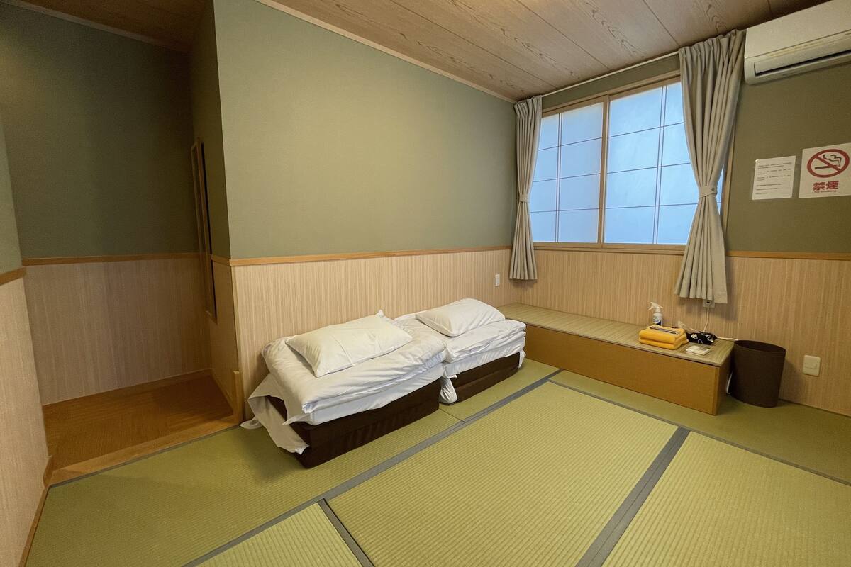 A room with a bed, a window with curtains, a bedside table with a bottle, and a wall-mounted air conditioner.