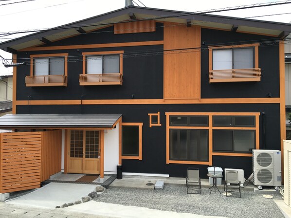 Une maison moderne à deux étages, avec une façade noire et des éléments décoratifs en bois, comprenant une porte coulissante, un petit porche avec des chaises et une grande fenêtre.