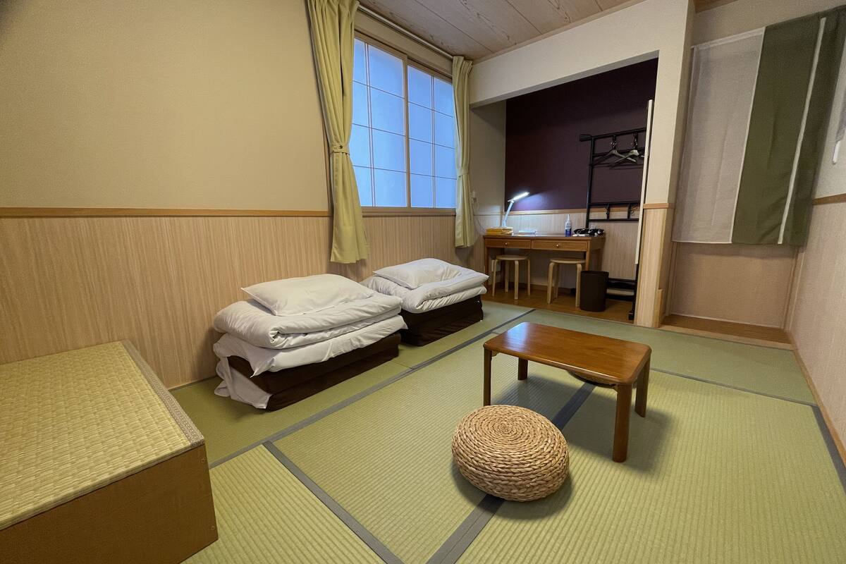 A traditional Japanese-style room with tatami mats, two single beds, a wooden table, a woven stool, and a desk with a lamp.