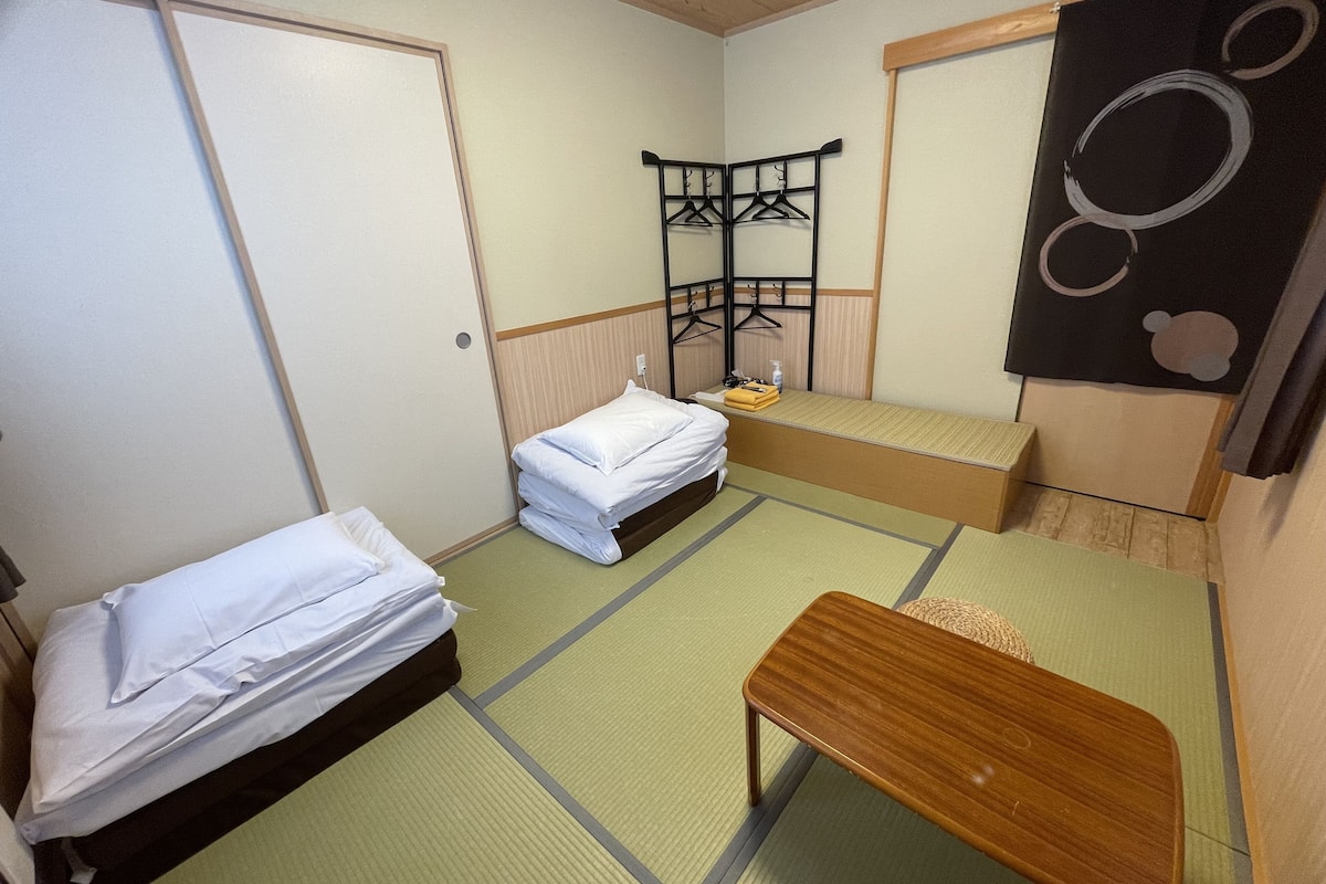 A compact hotel room with two beds, a wooden bench, a small table, and a wall-mounted shelf with hangers.