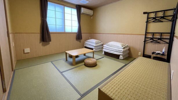 A traditional Japanese room with tatami flooring, a low wooden table, a futon bed, and a hanging clothes rack. - Guesthouse OC (Fujikawaguchiko)