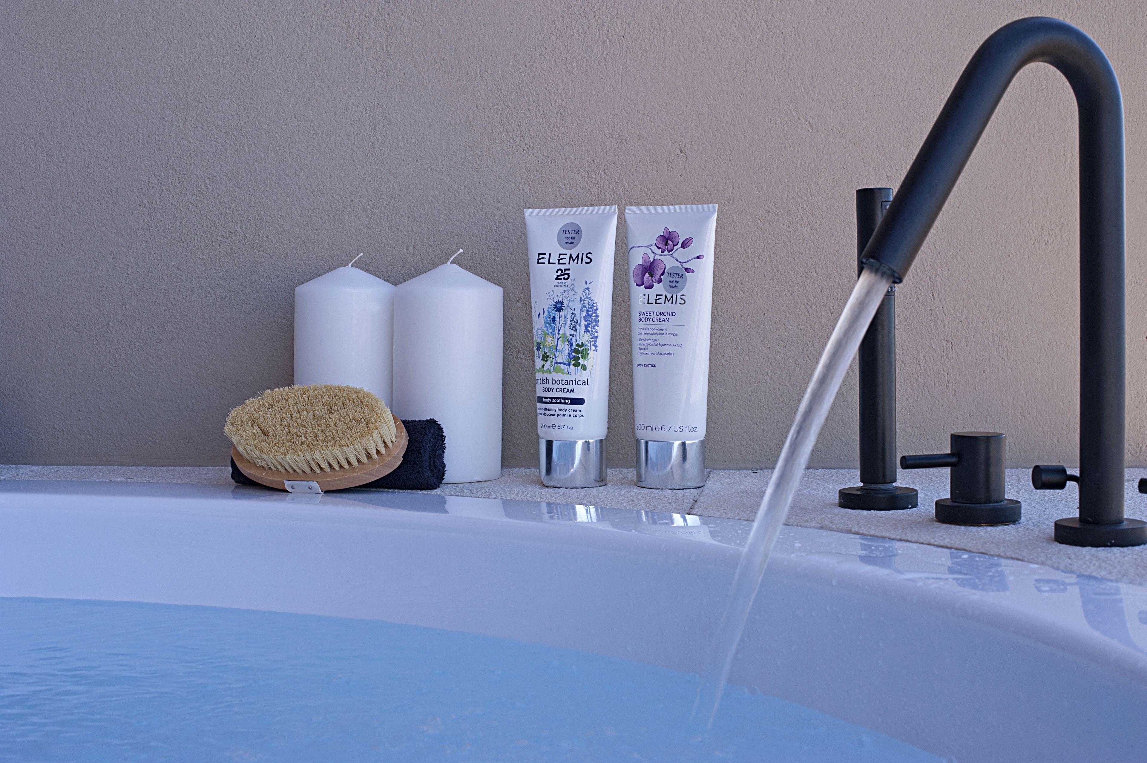 A bathroom with a bathtub, a hairbrush, two bottles of Elemis body cream, and a faucet.