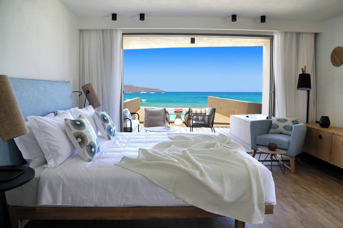 Ein Hotelzimmer mit einem großen Bett, einem Balkon mit Meerblick und einer Sitzecke mit Tisch und Stühlen.