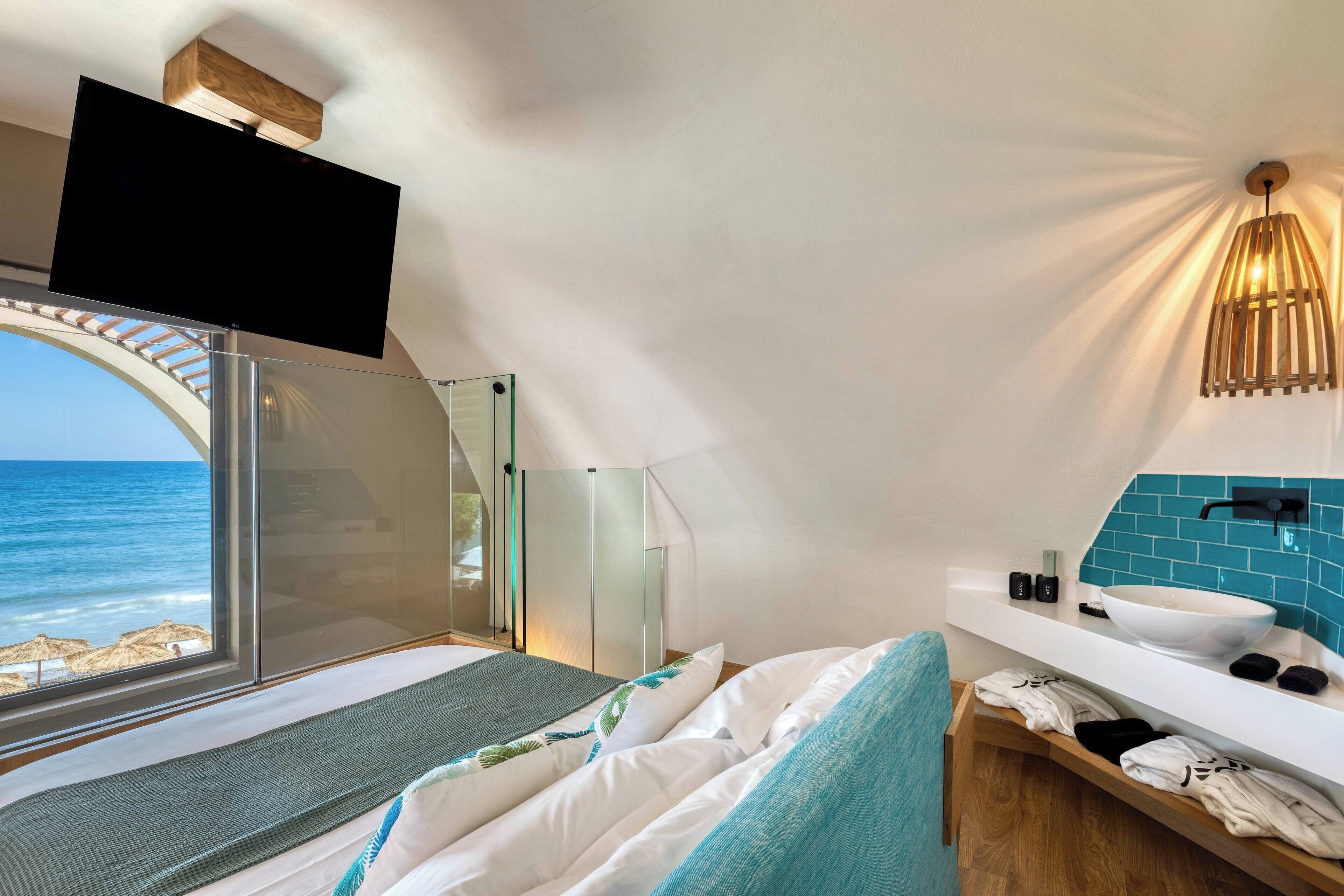 Un dormitorio moderno con un ventanal grande, un televisor de pantalla plana, techo de madera y un baño con lavamanos y perchero para toallas.