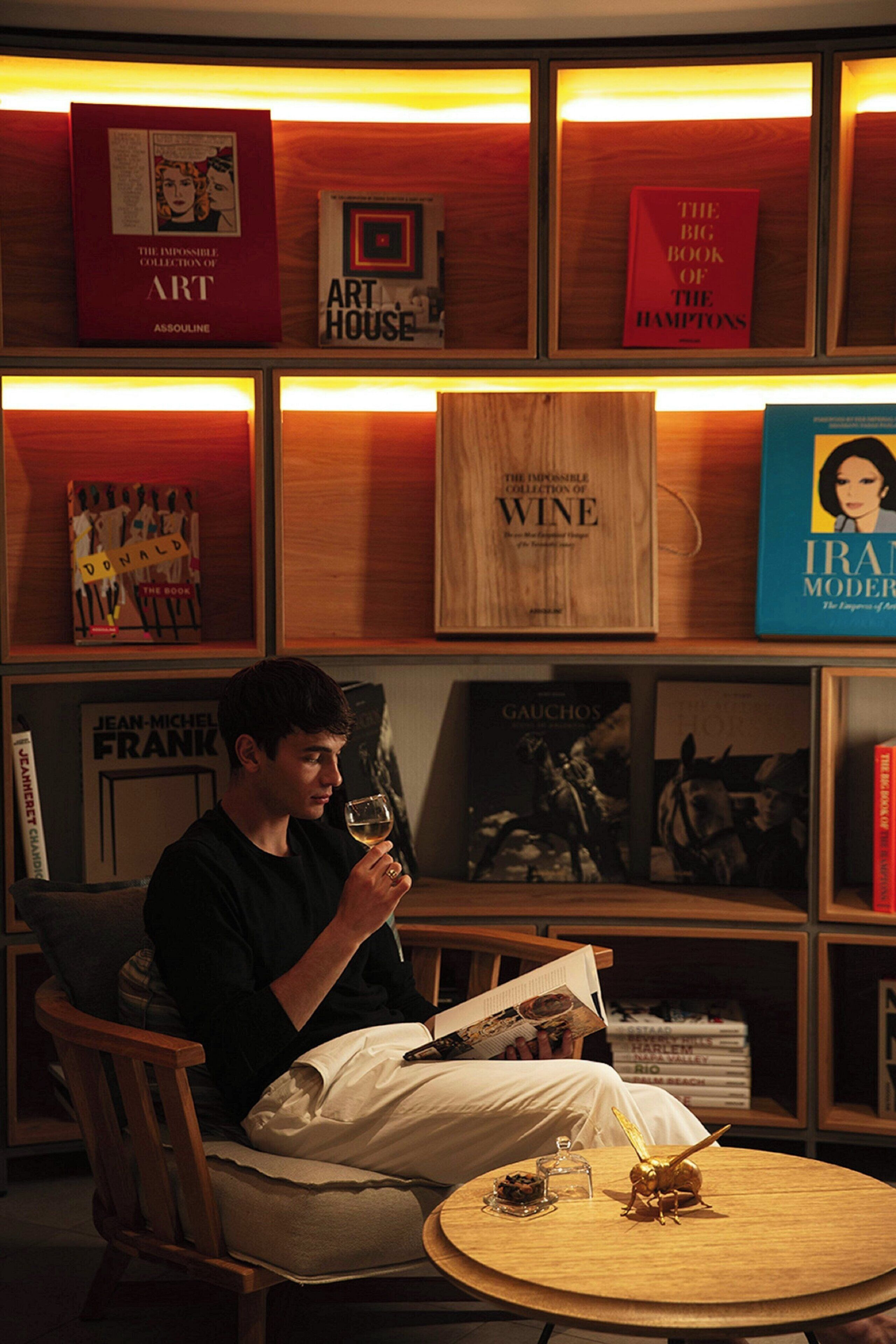 A person seated in a chair, reading a book, with a round wooden table in front. Shelves with books and framed pictures are visible in the background.