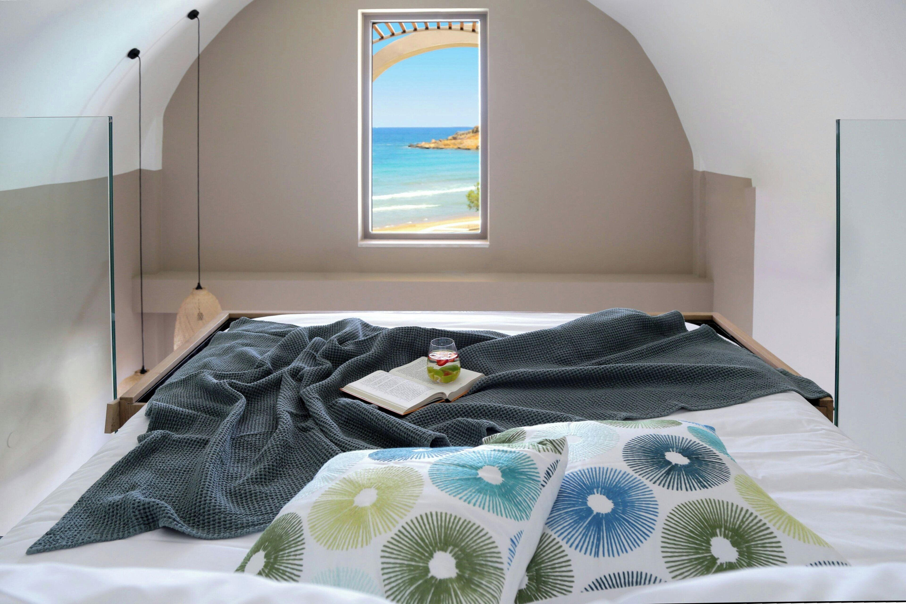 A bedroom with a bed, a window with a view of the sea, a book, and a glass of water on the bed.
