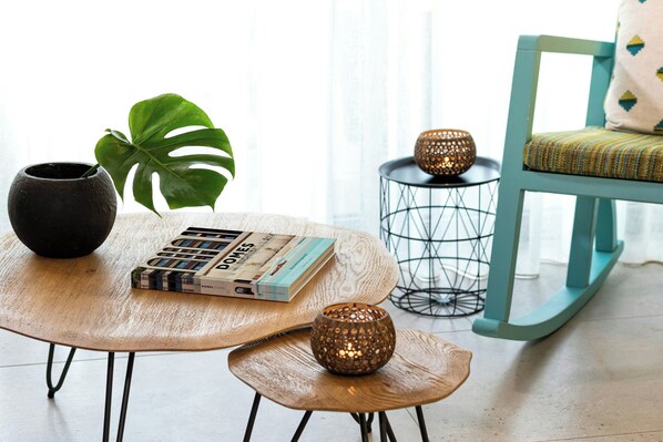 A cozy reading nook with a round wooden table, a black pot, a book, a small wooden stool, a green leafy plant, and a blue rocking chair.