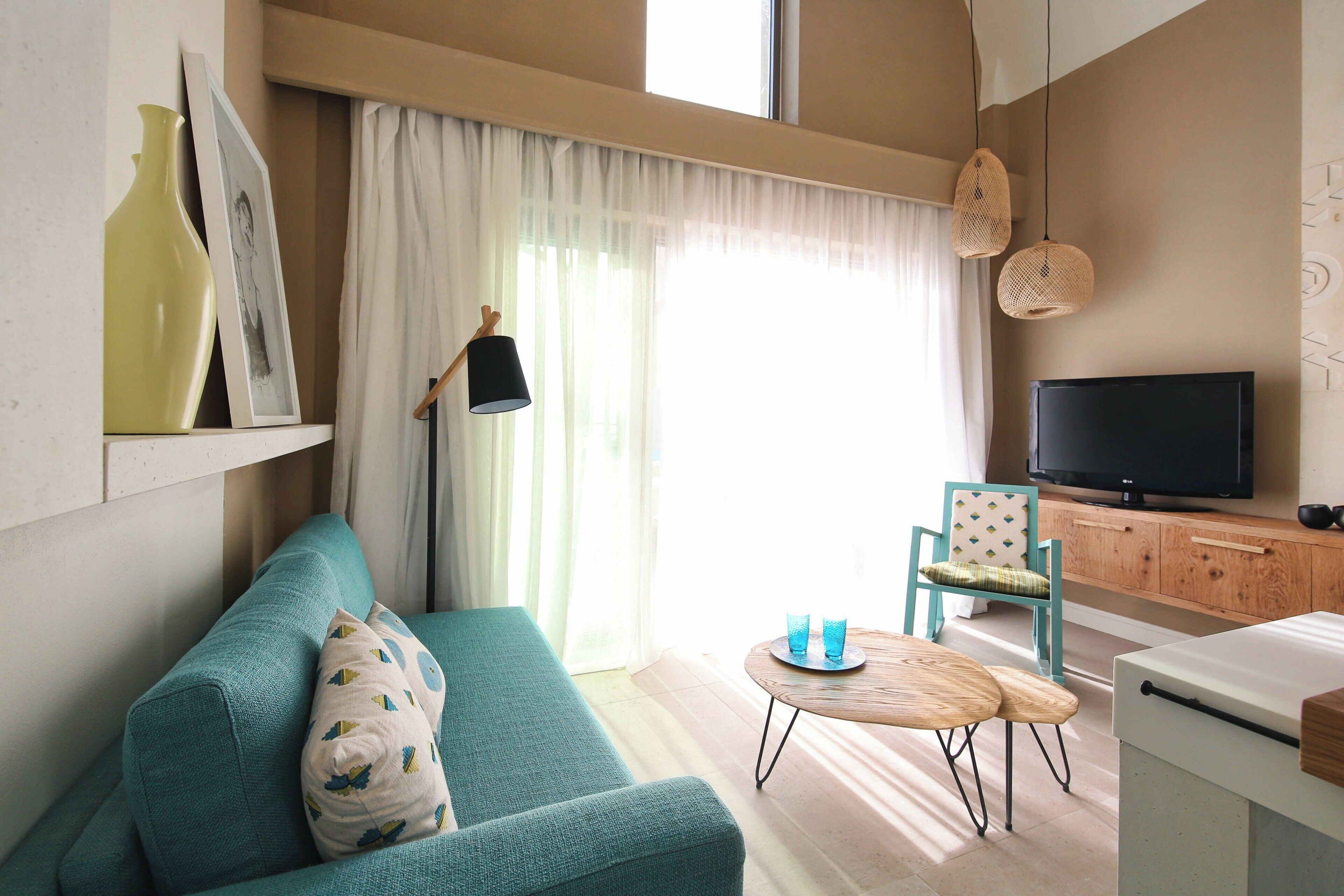 Una sala de estar con un sofá azul verdoso, una mesita de madera, un televisor y una ventana con cortinas de gasa.