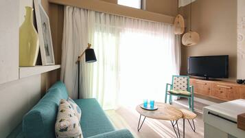 A living room with a teal sofa, a small wooden table, a television, and a window with sheer curtains.