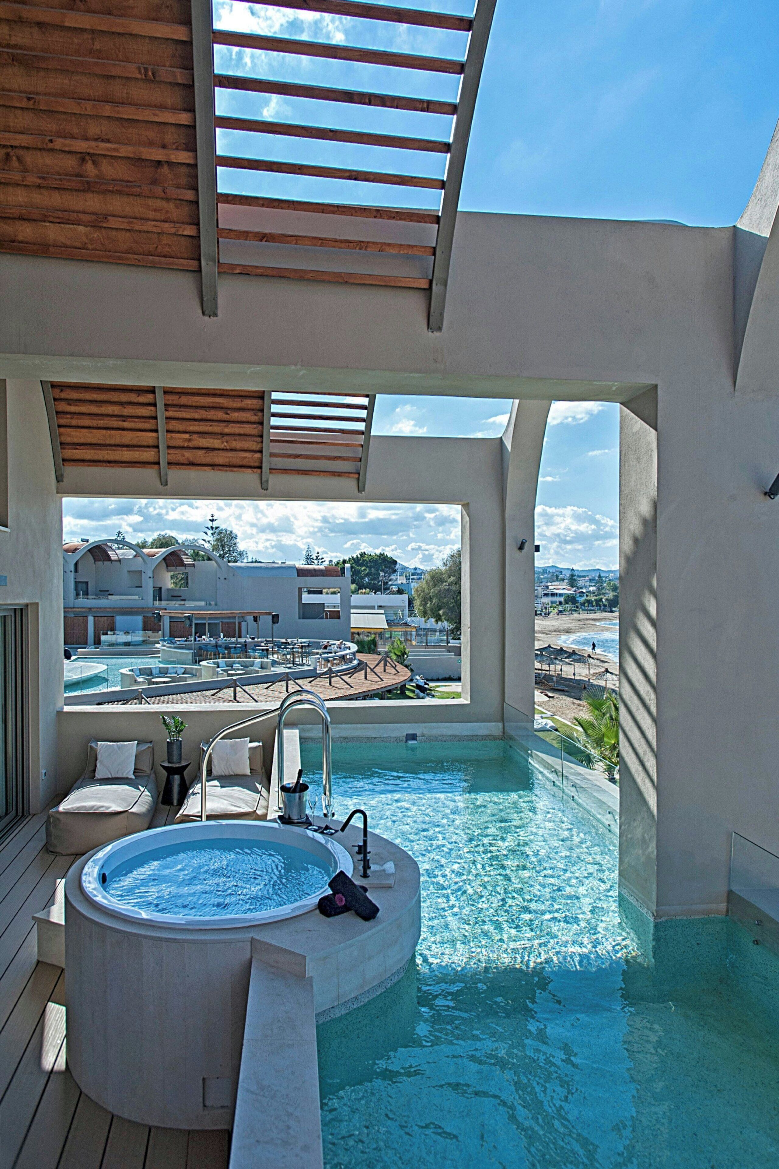 Un bain à remous avec vue sur une piscine et une plage, une terrasse en bois et un espace couvert agrémenté d’une pergola.