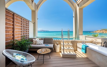 A sunlit outdoor lounge area with a sofa, chair, and small table, overlooking a clear blue sea and a distant island.