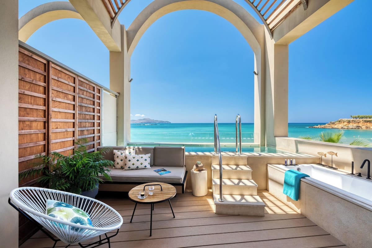 Eine sonnendurchflutete Außenlounge mit einer Couch, einem Sessel und einem kleinen Tisch mit Blick auf das klare, blaue Meer und eine entfernte Insel.