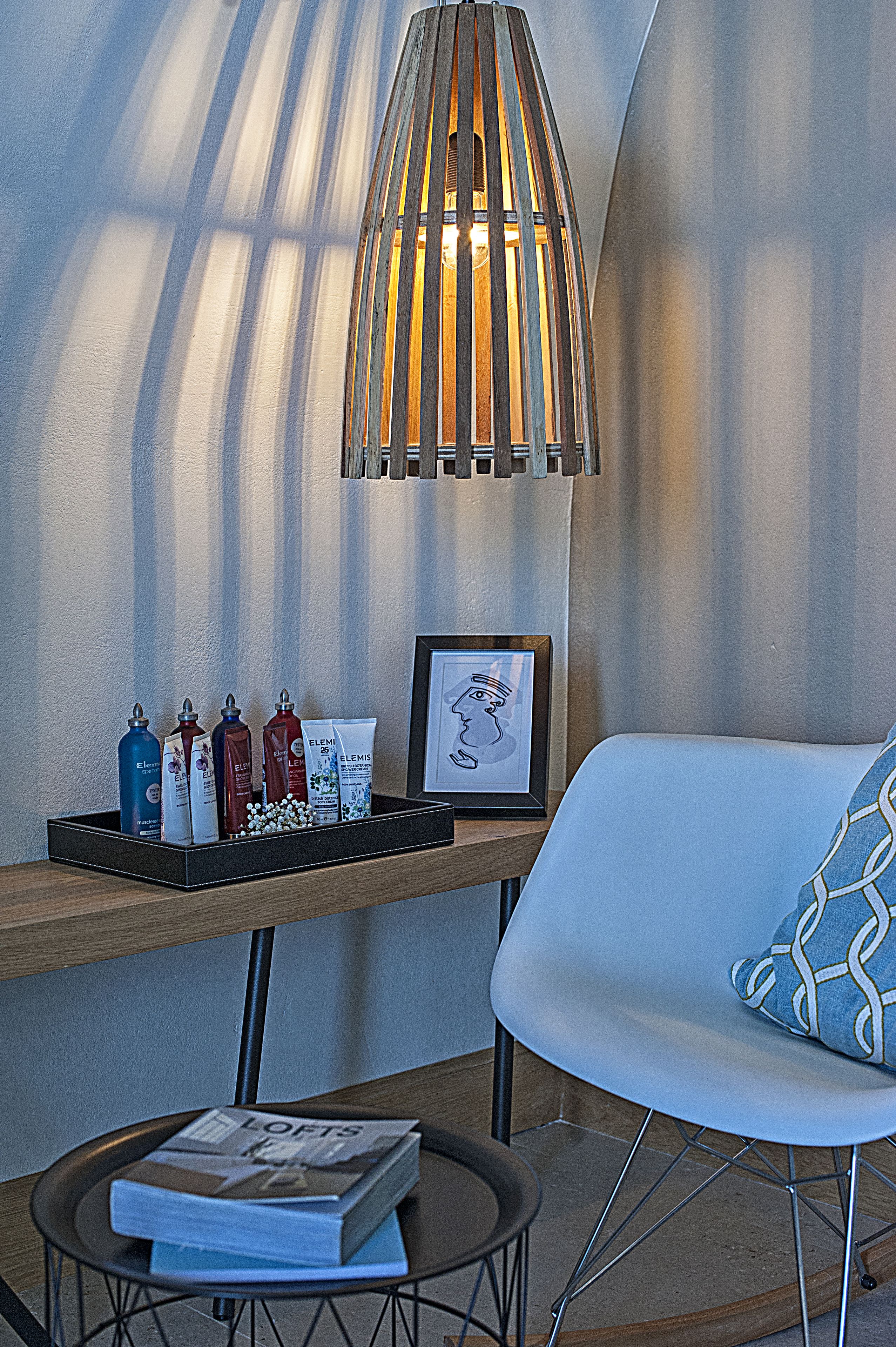 Una habitación moderna con una mesita de madera que sostiene artículos de aseo, un cuadro enmarcado y una revista. Una silla blanca con un cojín estampado está apoyada en la pared.