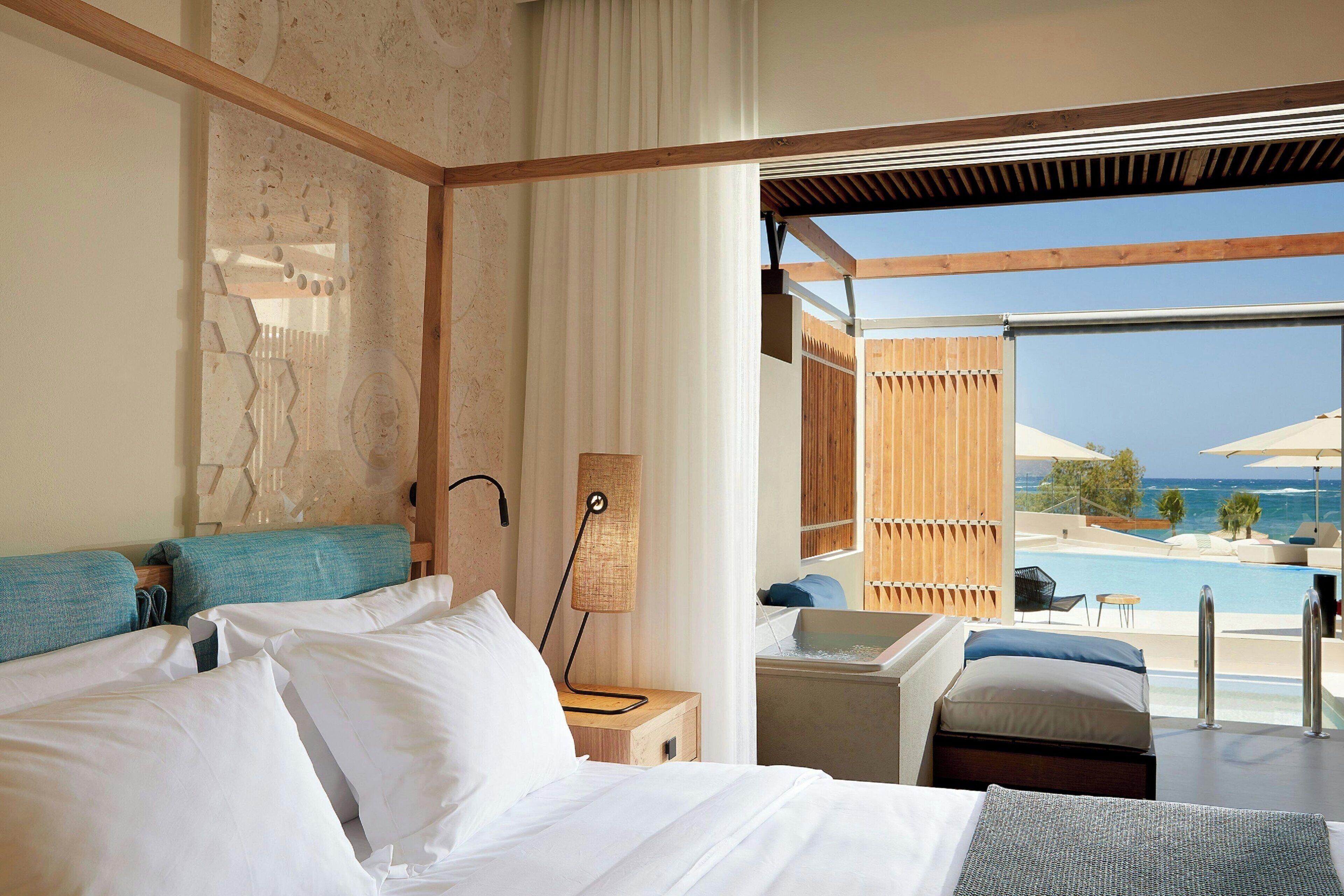 A hotel room with a bed, a bedside table, a lamp, and a view of a pool and ocean.
