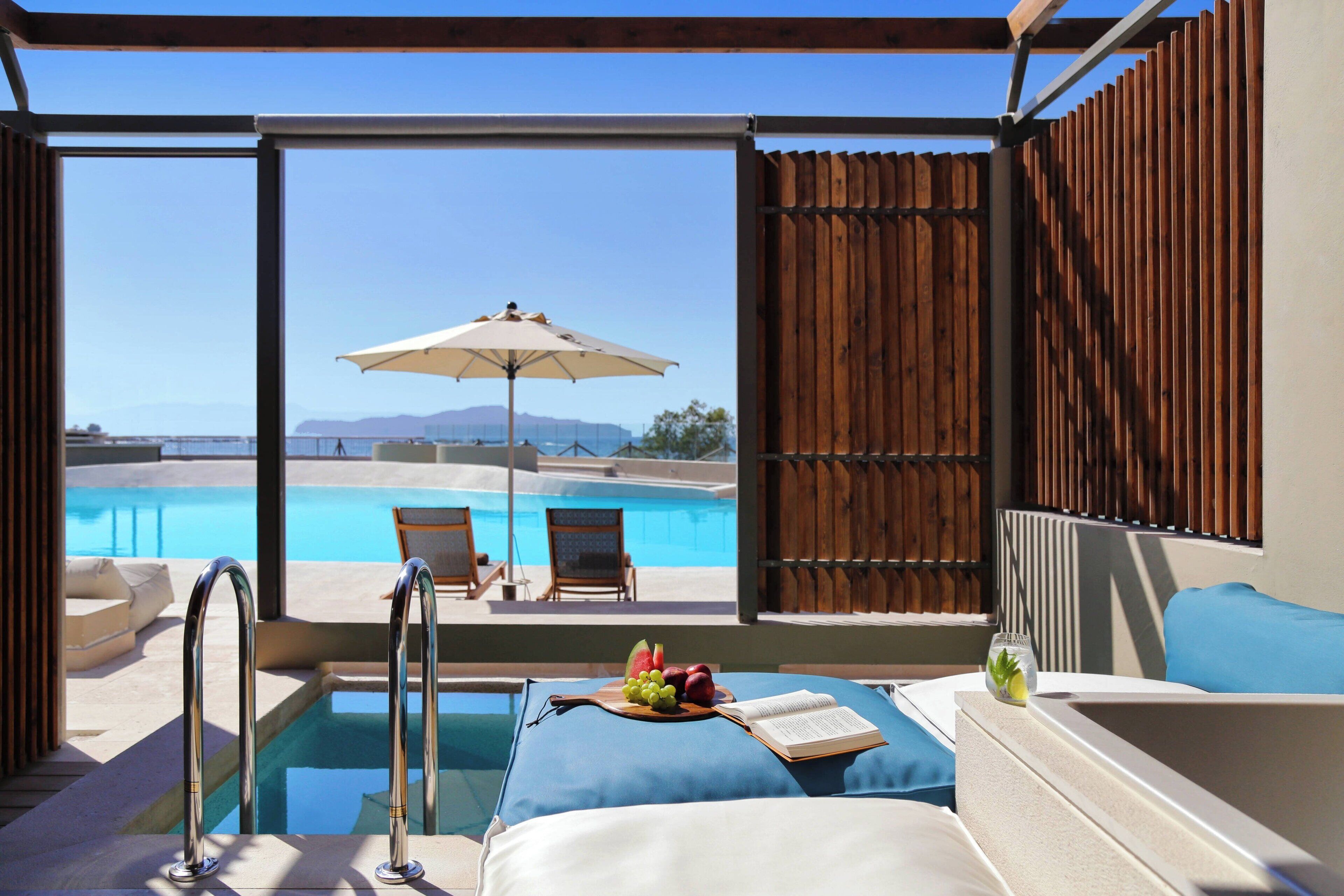 A poolside lounge with a view of a swimming pool, a wooden fence, and a sun umbrella.