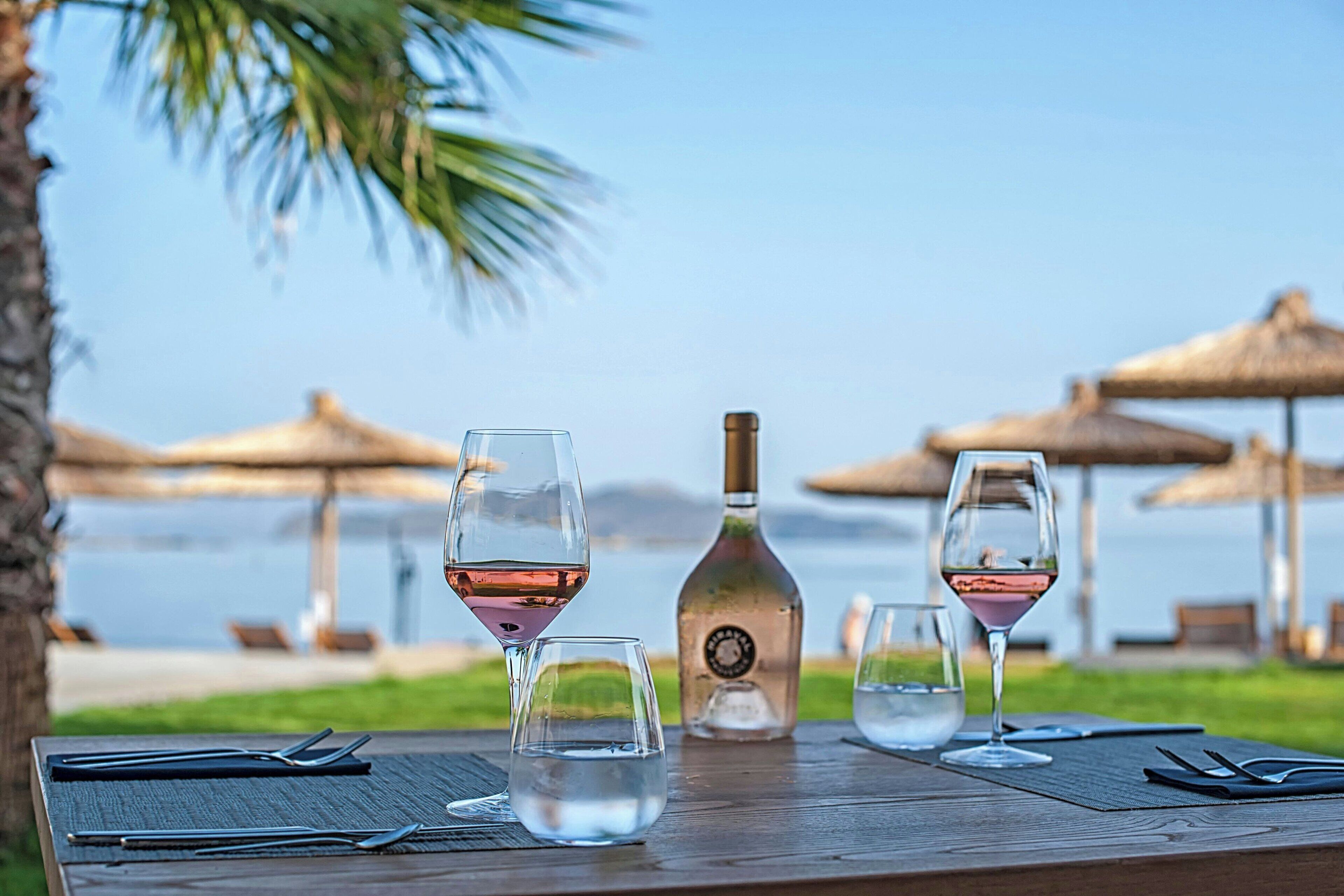 Una mesa de madera con copas de vino, una botella y cubiertos, frente a un paisaje con sombrillas y un cuerpo de agua.
