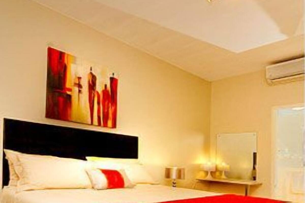 A hotel room with a large bed, a red throw blanket, a painting on the wall, and a nightstand with a lamp.