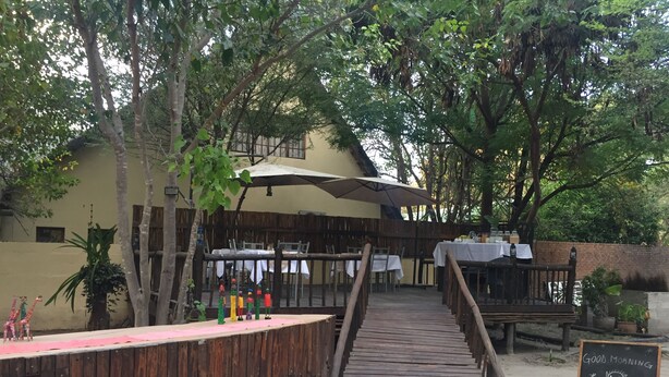 A wooden deck leading to a restaurant with outdoor seating, surrounded by trees and a covered area with umbrellas. - Peo Luxury Apartments (Maun)