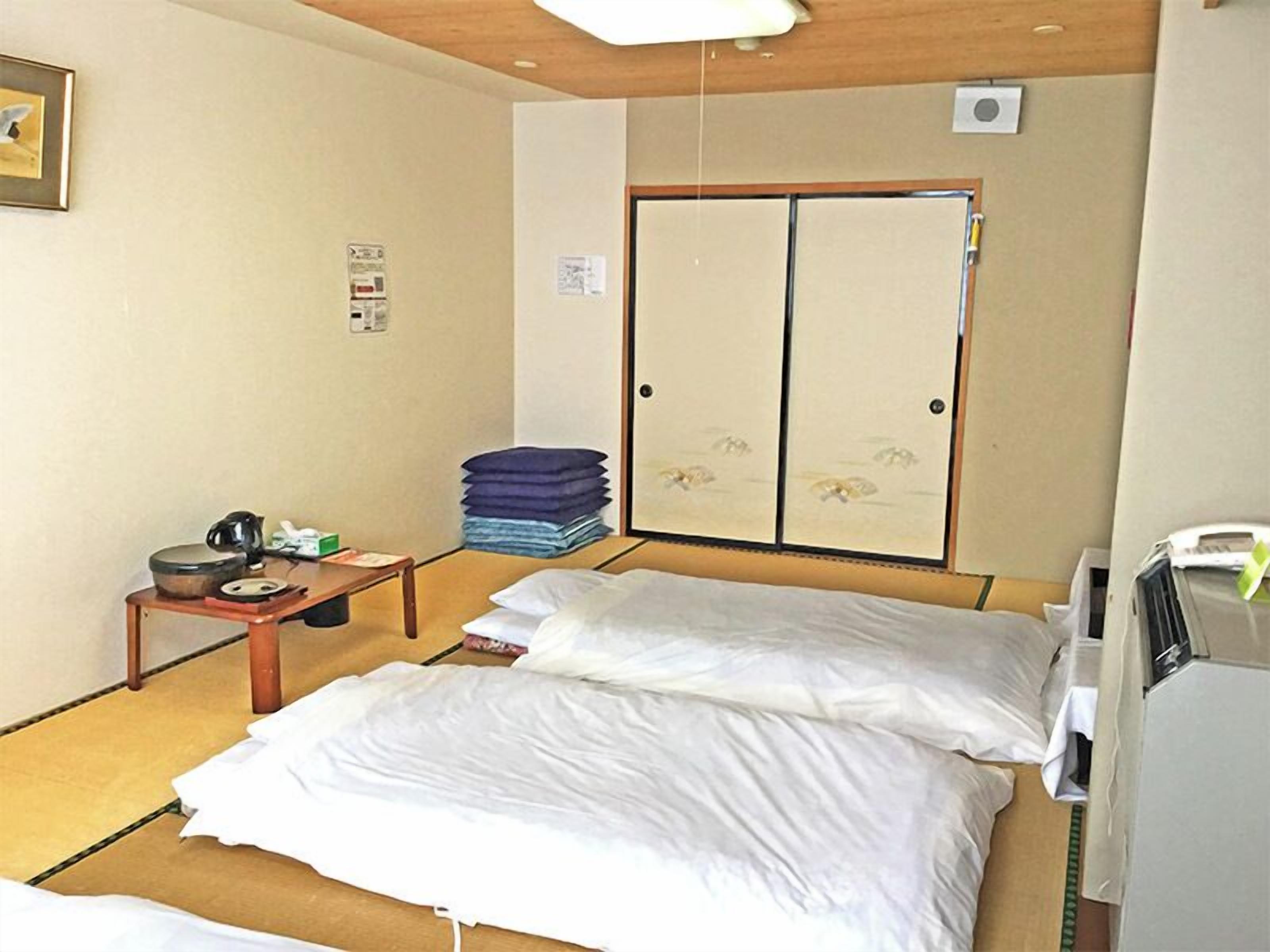 Een hotelkamer met een bed, een klein tafeltje met een waterkoker, een stapel opgevouwen kleding en een kledingkast.