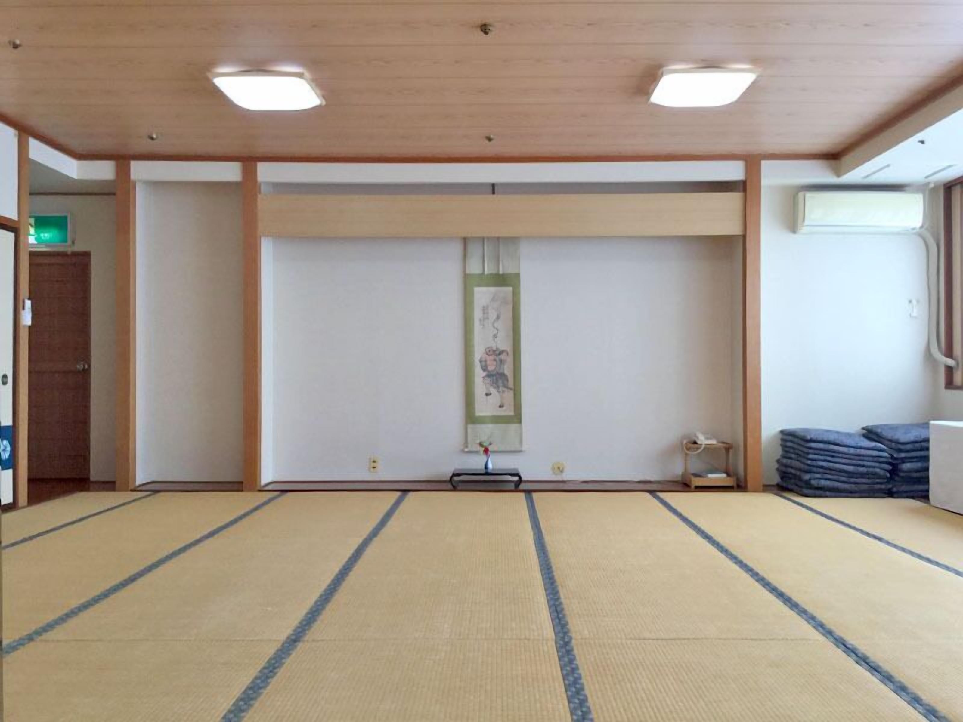 A tatami mat room with a wooden ceiling, a framed picture on the wall, and a stack of folded mats to the right.