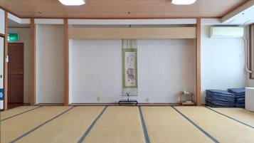 A tatami mat room with a wooden ceiling, a framed picture on the wall, and a stack of folded mats to the right.