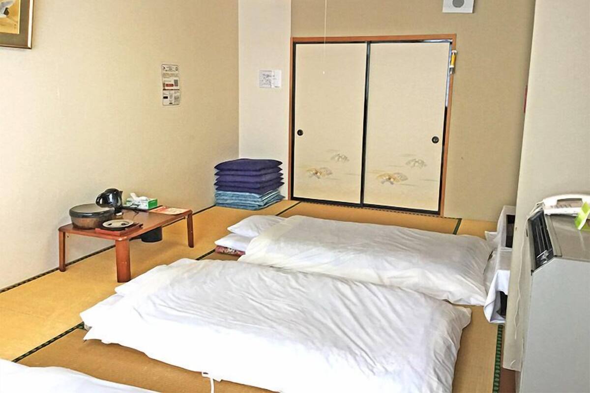 A hotel room with a bed, a small table with a kettle, a stack of folded clothes, and a wardrobe with a sliding door.