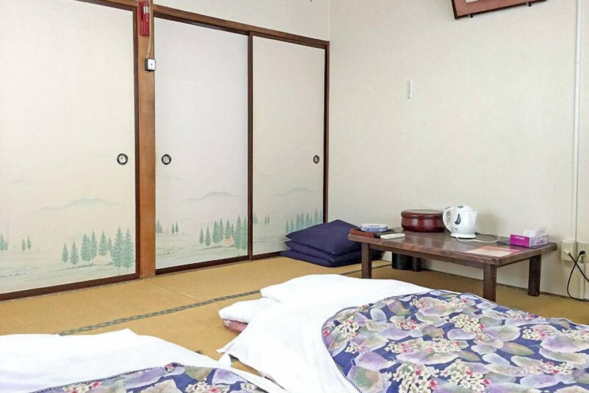A traditional Japanese room with tatami flooring, a low table with a kettle, and a bed with a floral bedspread.