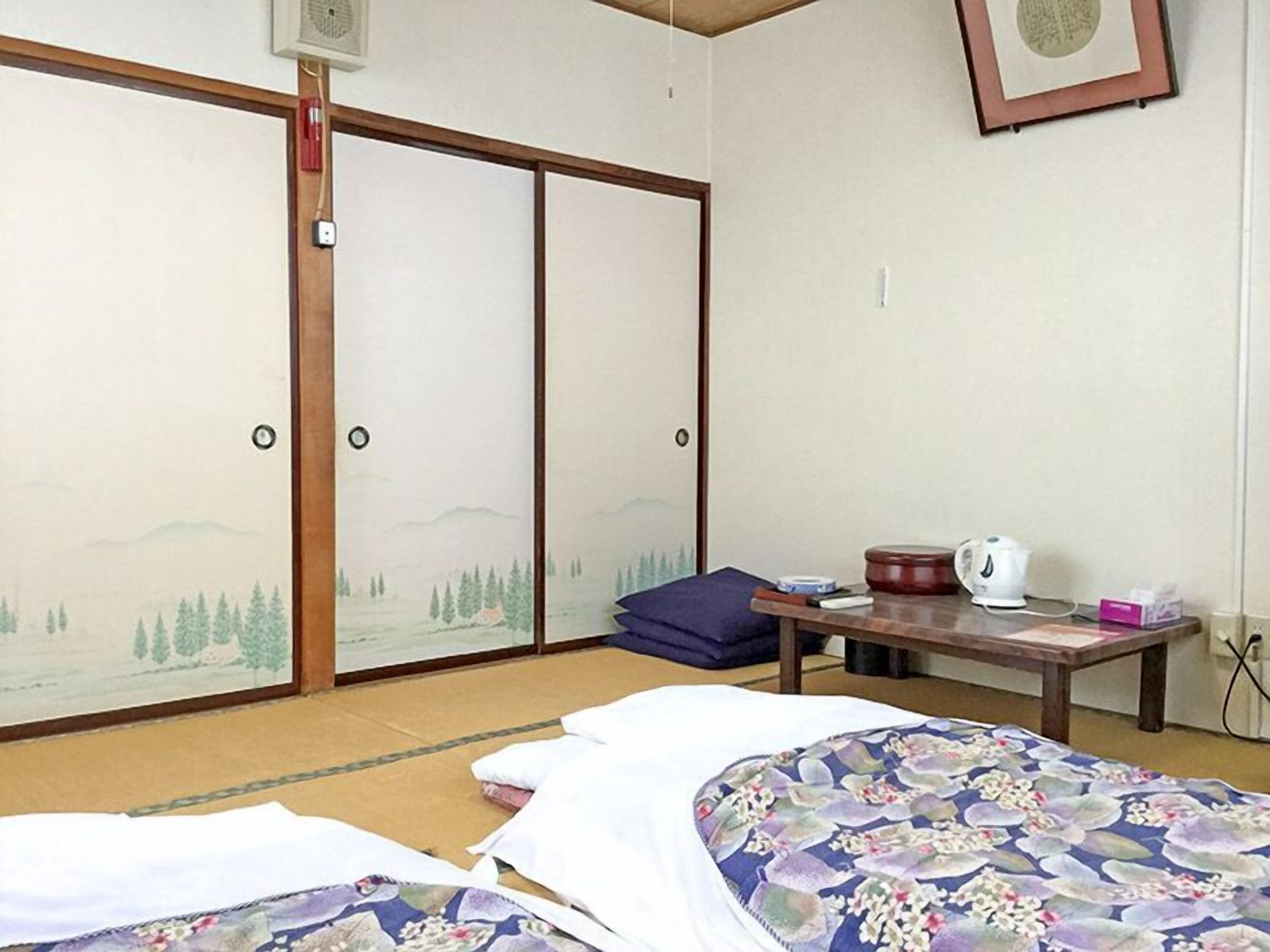 Een traditionele Japanse kamer met tatami vloeren, een bed met bloemenbedekking, een tafel met waterkoker en schuifdeuren met uitzicht op een landschap.