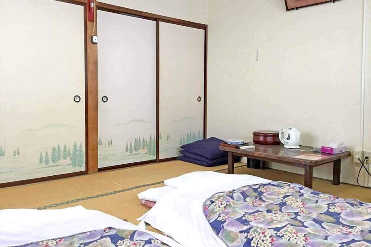 A traditional Japanese room with tatami flooring, a bed with a floral bedspread, a table with a kettle, and sliding doors with a landscape view.