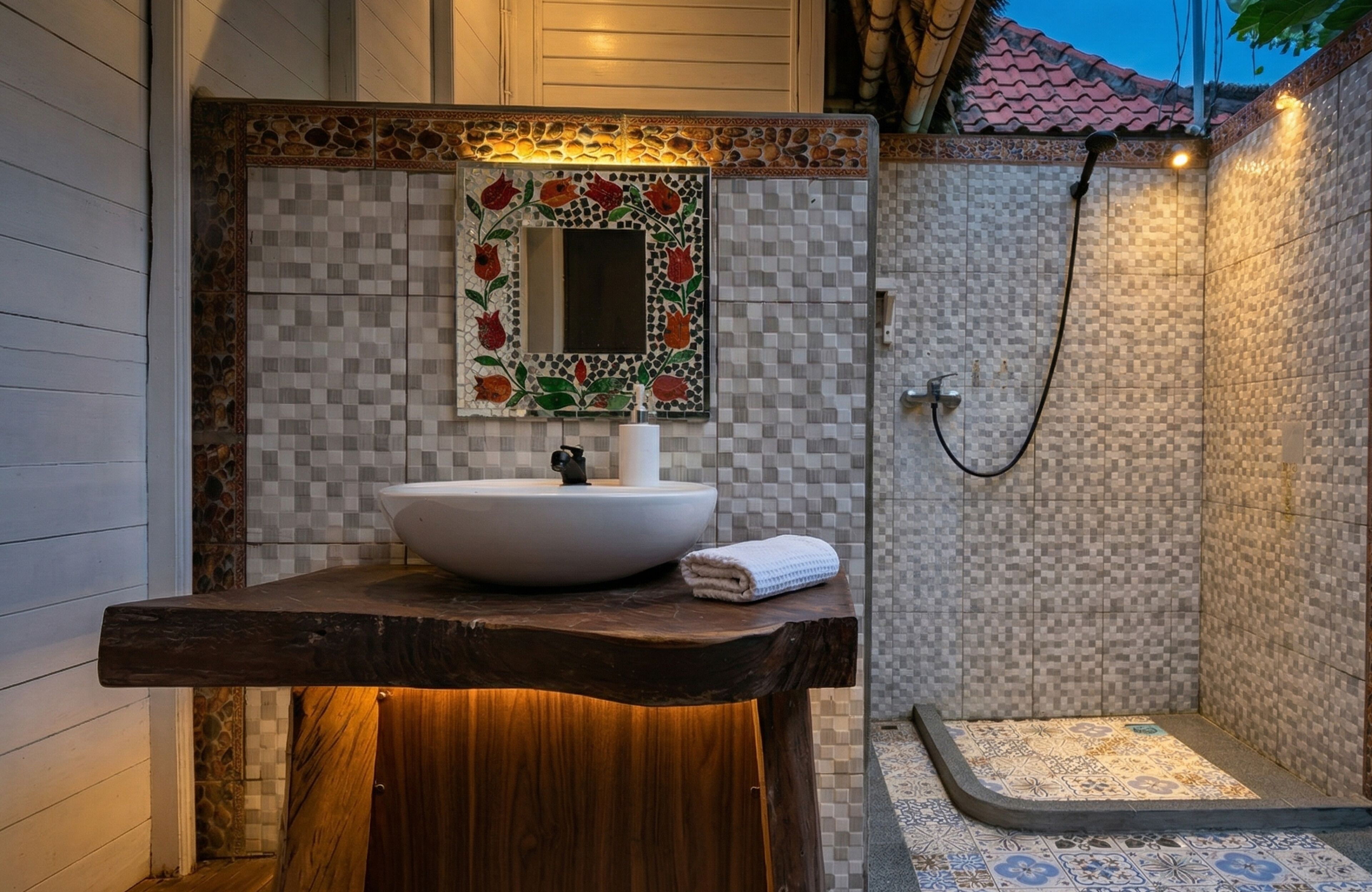 A bathroom with a wooden vanity, a white sink, a mirror with floral tile, and a shower area.