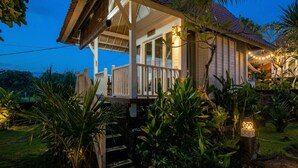 Een huis met een veranda, omgeven door tropische planten en een klein waterpartij.