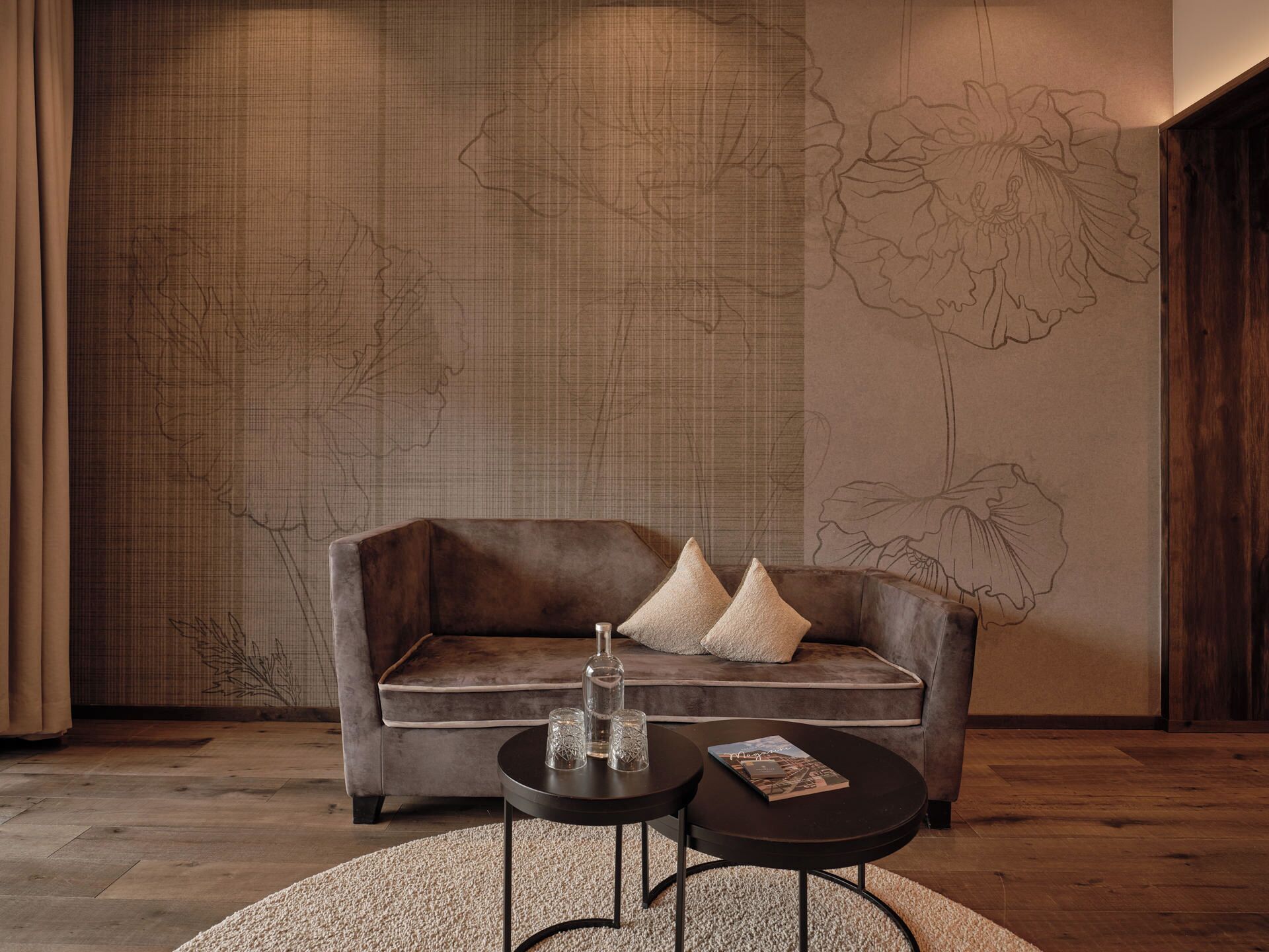Un salon moderne avec un canapé gris, deux tables d’appoint noires et un mur décoratif orné d’un motif floral.