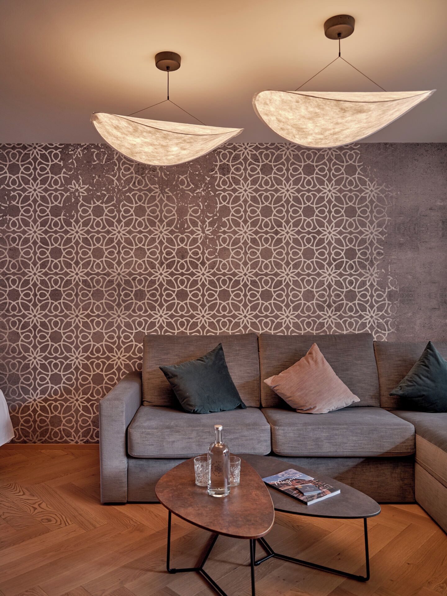 A modern living room with a patterned wallpaper, a grey sofa with cushions, a coffee table with a bottle and glasses, and two pendant lights.