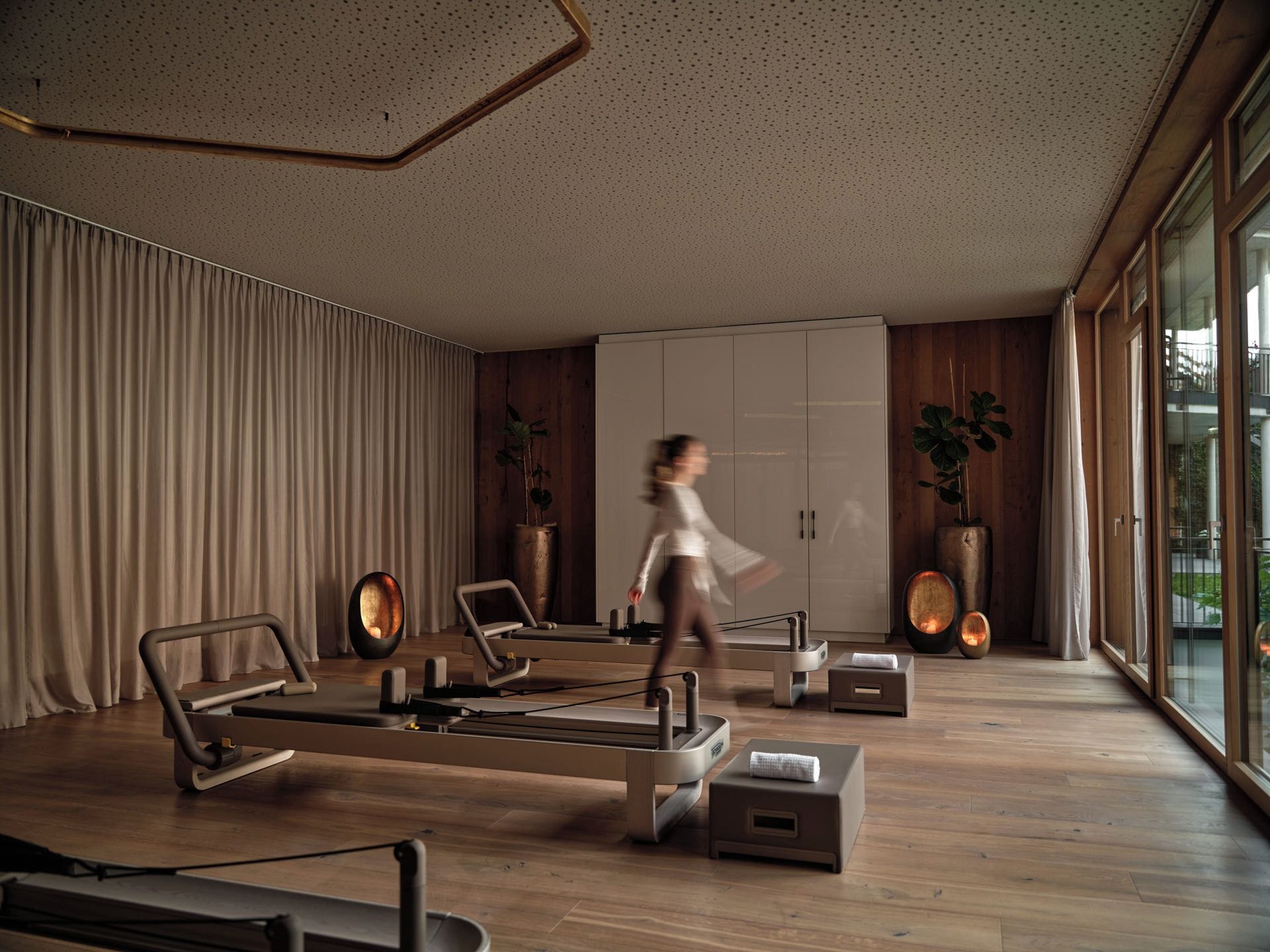 A modern living room with a Pilates reformer, a large wardrobe, and a view of the outdoors through large windows.