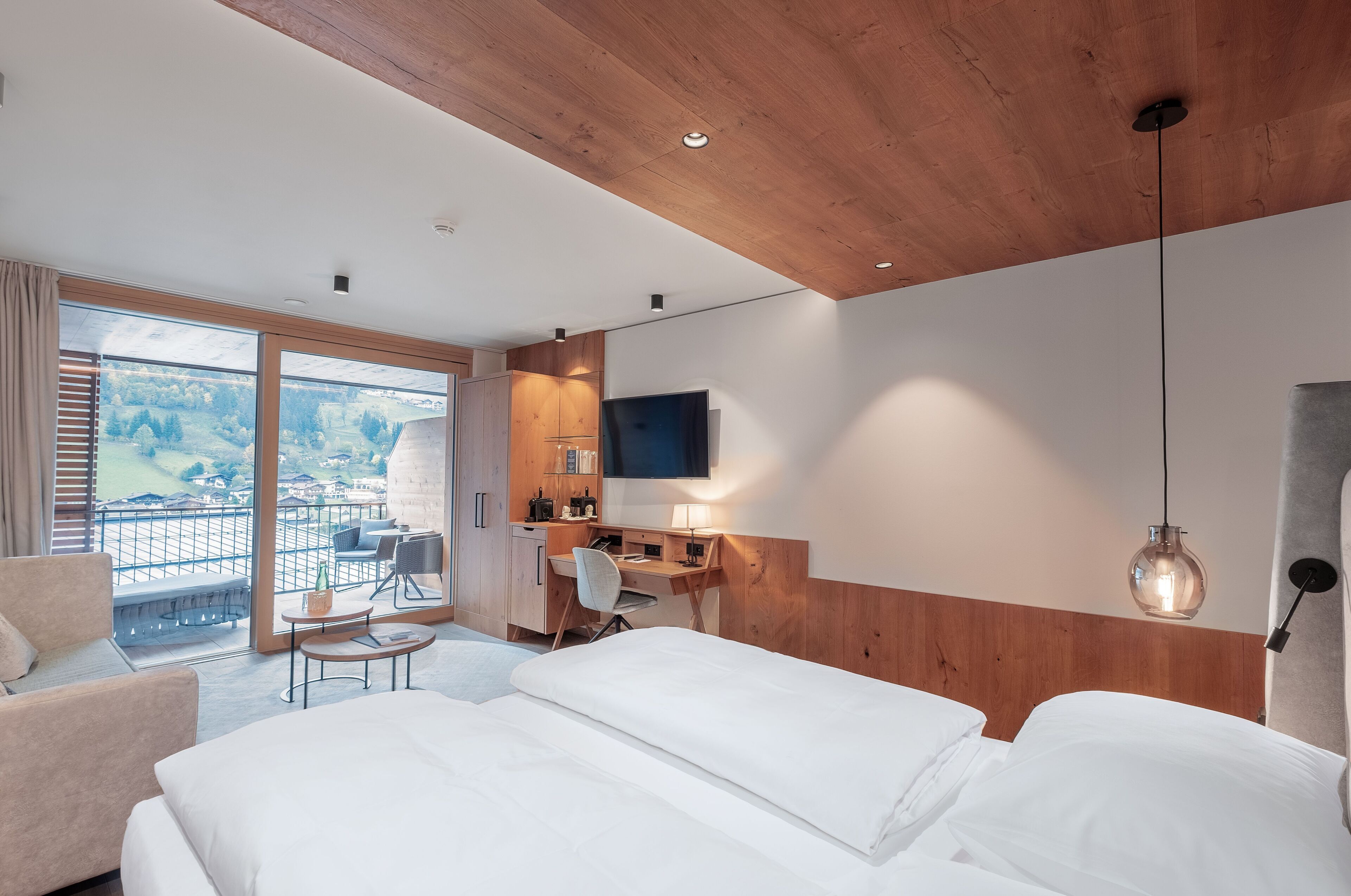 Ein Hotelzimmer mit einem großen Bett, einem Schreibtisch, einem Sessel, einem Fernseher und einem Balkon mit Blick auf die Landschaft.
