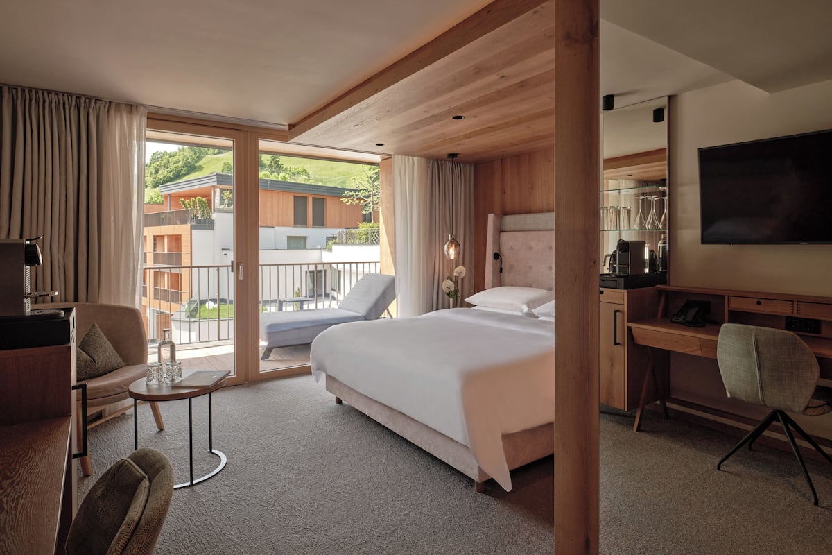 Ein modernes Hotelzimmer mit einem großen Bett, einem Schreibtisch, einem Sessel und einem Balkon mit Blick auf die umliegenden Gebäude.