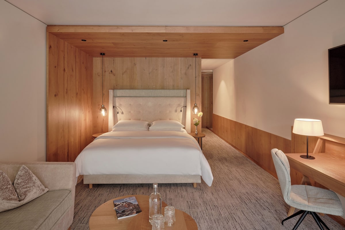 Ein Hotelzimmer mit einem großen Bett, einem Holzkopfbrett, einem kleinen runden Tisch mit einer Zeitschrift und Gläsern, einem Sessel und einem Schreibtisch mit einer Lampe.